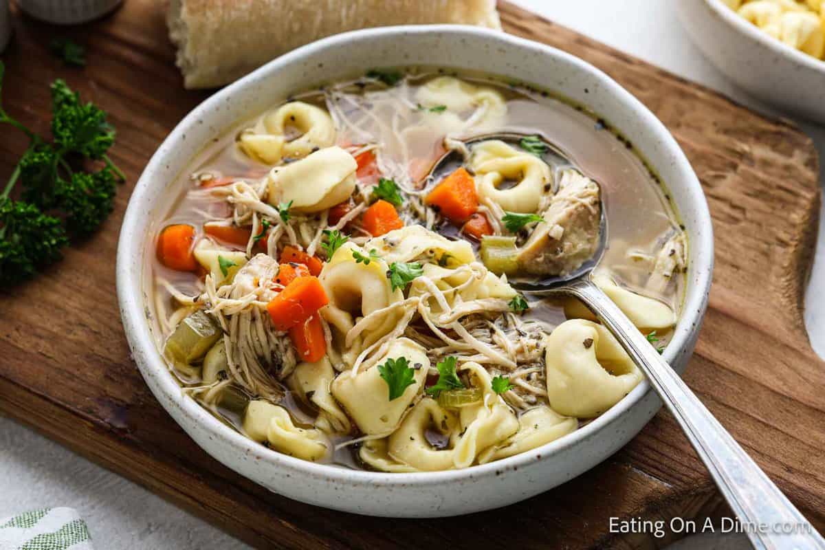 A bowl of crockpot chicken tortellini soup with carrots, celery, and a parsley garnish. A spoon rests in the comforting soup, while a piece of bread sits invitingly on a wooden cutting board beside the bowl.