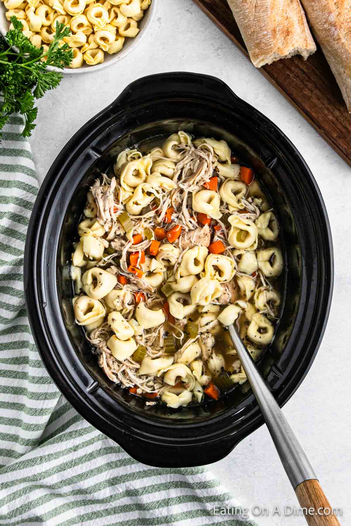 A crockpot brimming with chicken tortellini soup showcases pasta, tender shredded chicken, and diced carrots swimming in a savory broth. Nearby, fresh bread awaits next to a bowl of uncooked tortellini, while a green-striped cloth adds a touch of charm.
