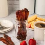 Candied bacon strips, made from a delicious candied bacon recipe, are arranged upright in a glass jar. In the background, waffles with strawberries, a cup of coffee, and white plates with forks rest on a light countertop.
