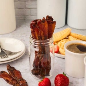 Candied bacon strips, made from a delicious candied bacon recipe, are arranged upright in a glass jar. In the background, waffles with strawberries, a cup of coffee, and white plates with forks rest on a light countertop.