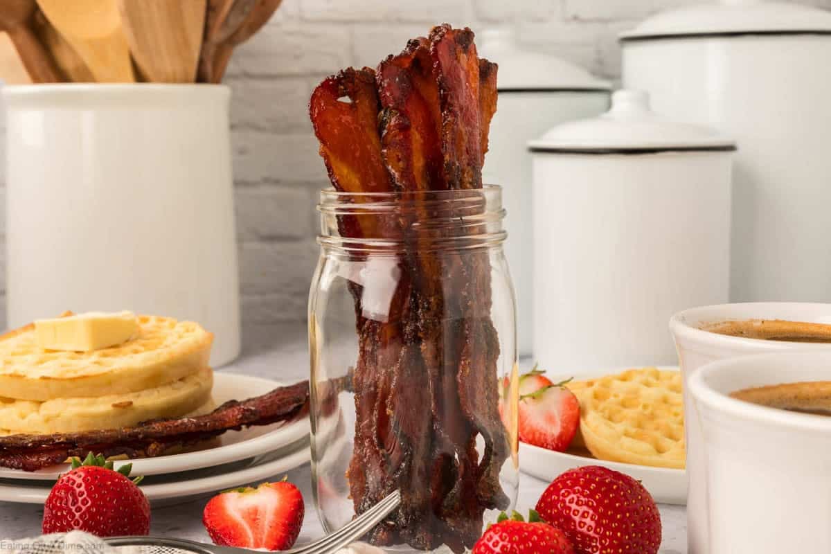A jar filled with crispy candied bacon strips sits on a breakfast table with waffles, strawberries, and mugs of coffee, alongside white canisters and kitchen utensils—perfect inspiration for your next candied bacon recipe.