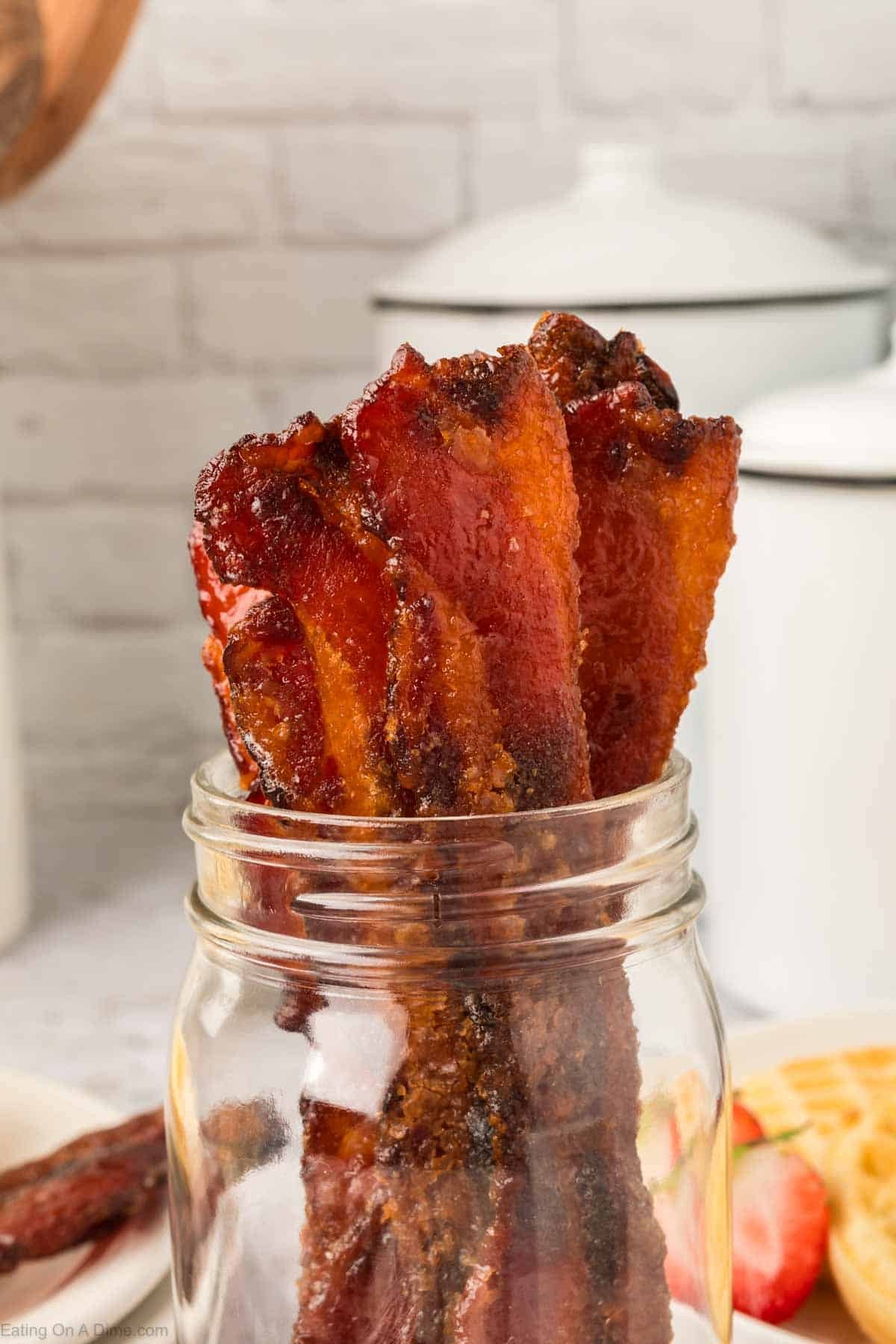 Several crispy, glazed bacon strips from a delicious candied bacon recipe are standing upright in a clear glass mason jar, with a light kitchen background and white canisters visible behind them.