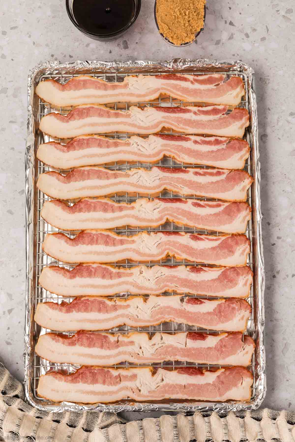 A baking sheet lined with foil and a wire rack holds twelve uncooked bacon strips in neat rows—perfect for a candied bacon recipe. Nearby, a bowl of brown sugar and a glass of dark liquid are visible on a light countertop.