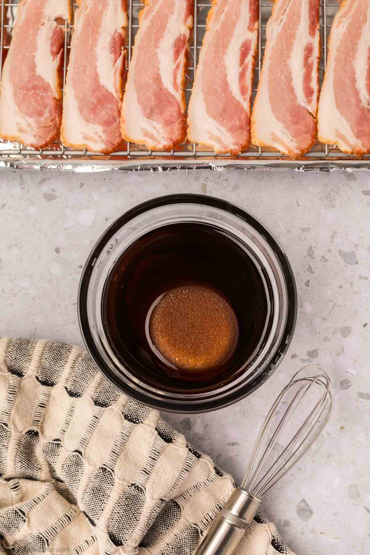 A bowl of dark liquid marinade with a whisk sits on a patterned towel; above, raw bacon strips—perfect for a candied bacon recipe—are arranged on a wire rack over a baking sheet.