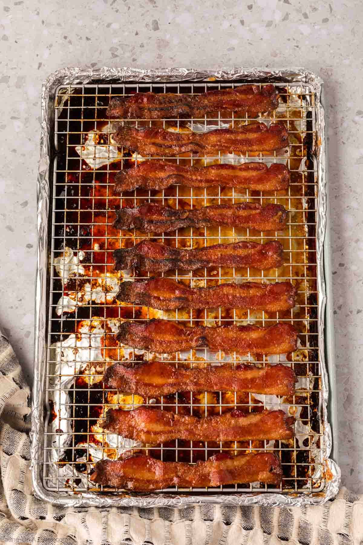 A baking tray lined with foil and a wire rack holds several strips of crispy bacon, perfect for a candied bacon recipe. The tray rests on a light countertop with a beige textured cloth partially visible in the corner.