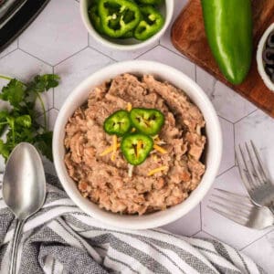 A bowl of Crockpot Refried Beans topped with shredded cheese and sliced jalapeños, surrounded by a spoon, forks, a striped napkin, cilantro, and bowls of sliced jalapeños and black olives.