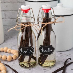 Two glass bottles with swing-top lids, each labeled "Vanilla" and filled with clear liquid and whole vanilla beans, show How to Make Vanilla Extract at Home. Twine is tied around the neck of each bottle as they sit on a light countertop.