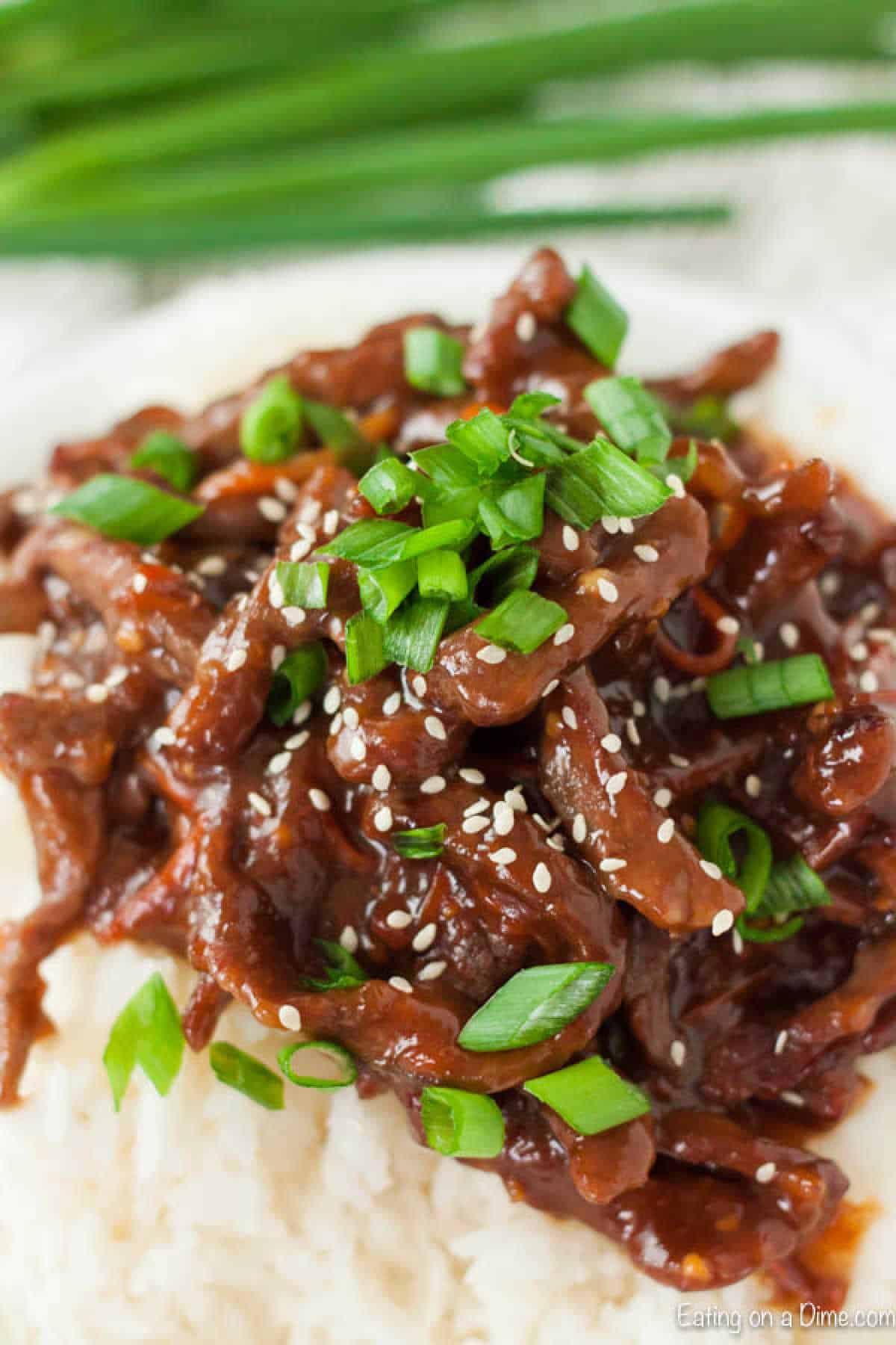 No need to get take out when you can make this easyย Slow Cooker Mongolian Beef. The flavors blend together for the best dish that your family will love. A plate of beef stir fry, reminiscent of slow cooker Mongolian beef, is garnished with sliced green onions and sesame seeds atop a bed of white rice. Green onion stalks are blurred in the background.