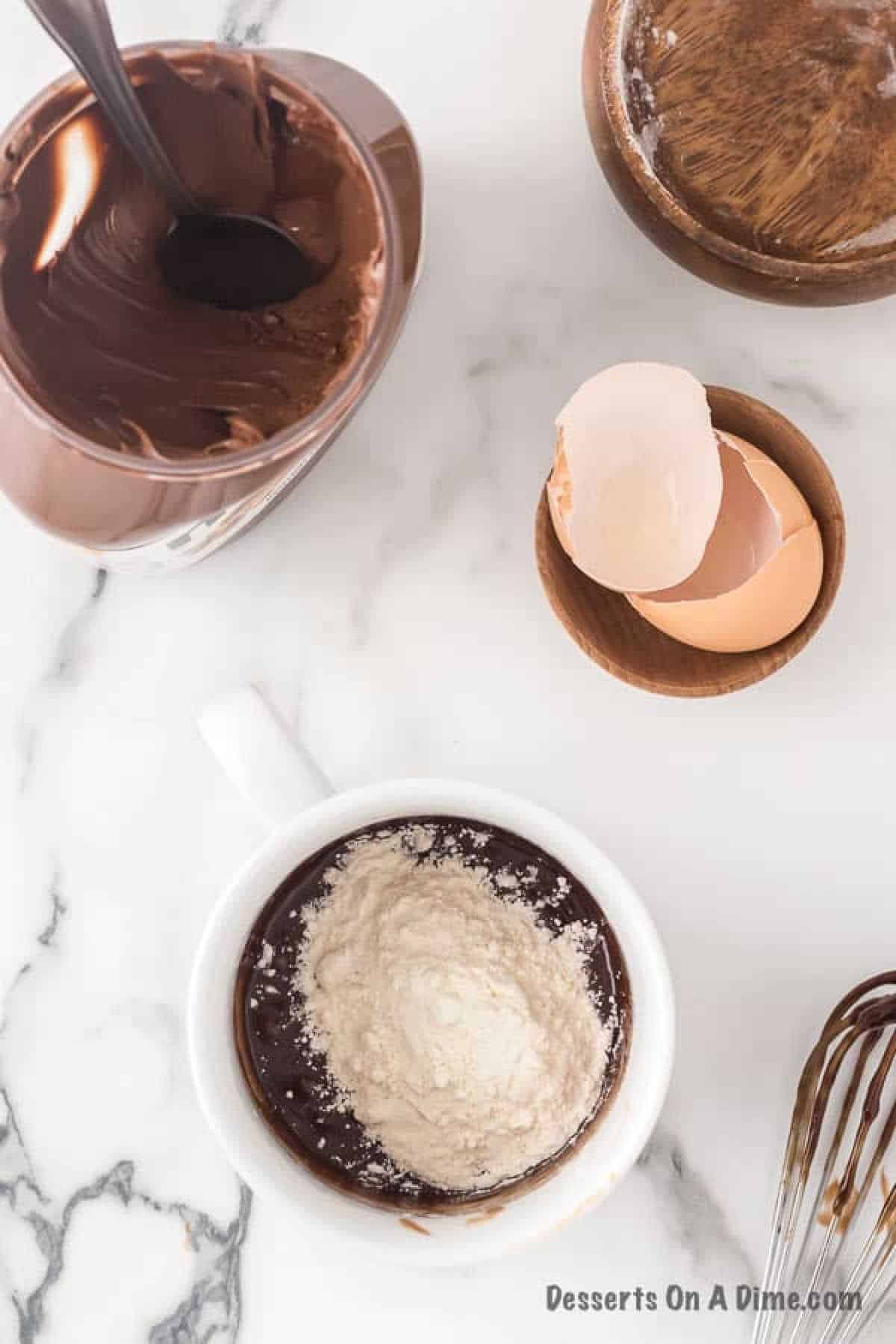 A mug filled with chocolate cake and topped with flour sits on a marble surface. Nearby, a jar of Nutella spread, perfectly poised for drizzling, accompanies a bowl with two eggshells and a whisk—essentials for crafting the ultimate Nutella mug cake.