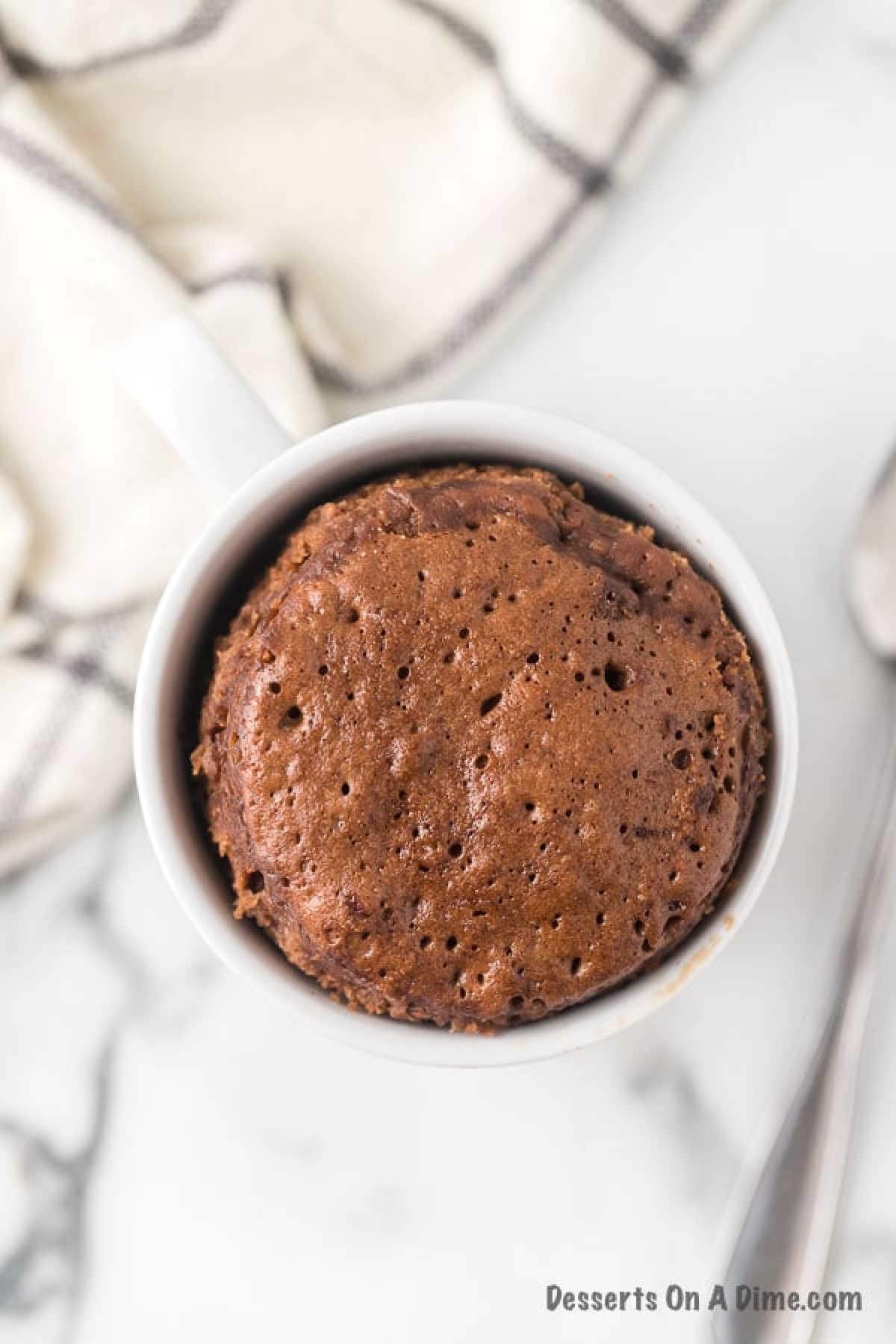 A Nutella mug cake sits in a white mug, with a cloth and spoon nearby. The cake's surface is dotted with small holes and slightly risen above the mug's rim, tempting any dessert lover. The image features a watermark that reads "Desserts On A Dime.com".