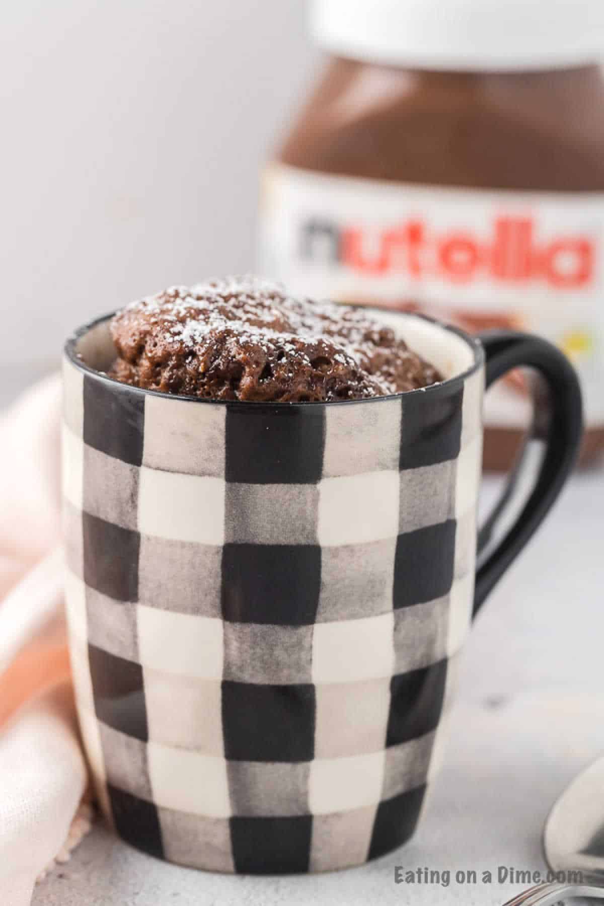 A black and white checkered mug cradles a Nutella mug cake, dusted with powdered sugar. A jar of the chocolate spread is blurred in the background, adding allure, while a soft pink cloth completes the cozy scene.