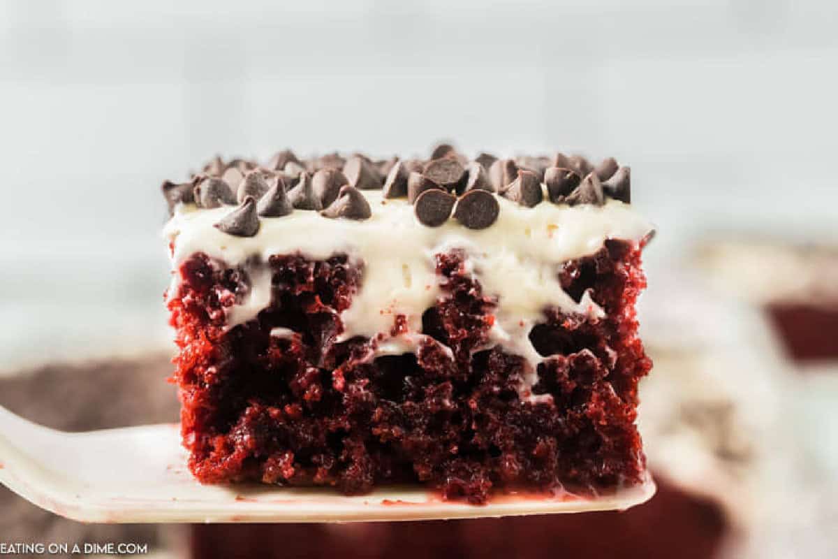 A close-up of a Red Velvet Poke Cake slice with white frosting and chocolate chips on top, held up on a spatula.