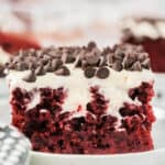 A close-up of a Red Velvet Poke Cake slice, topped with creamy white frosting and chocolate chips, served on a white plate.