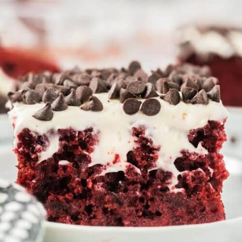 A close-up of a Red Velvet Poke Cake slice, topped with creamy white frosting and chocolate chips, served on a white plate.