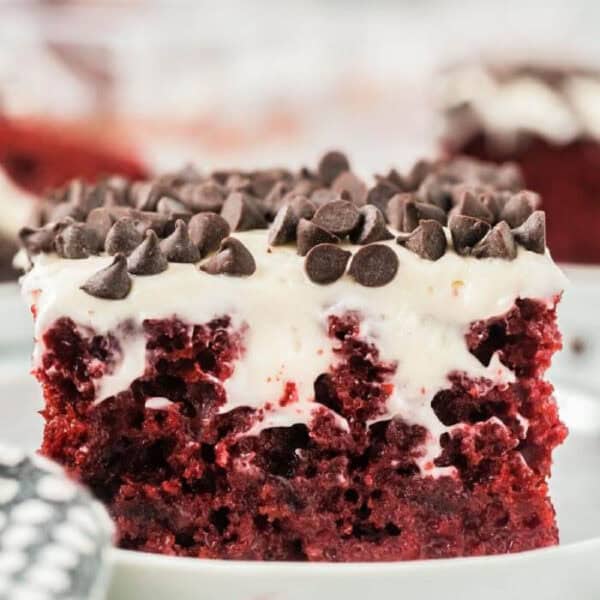 A close-up of a Red Velvet Poke Cake slice, topped with creamy white frosting and chocolate chips, served on a white plate.