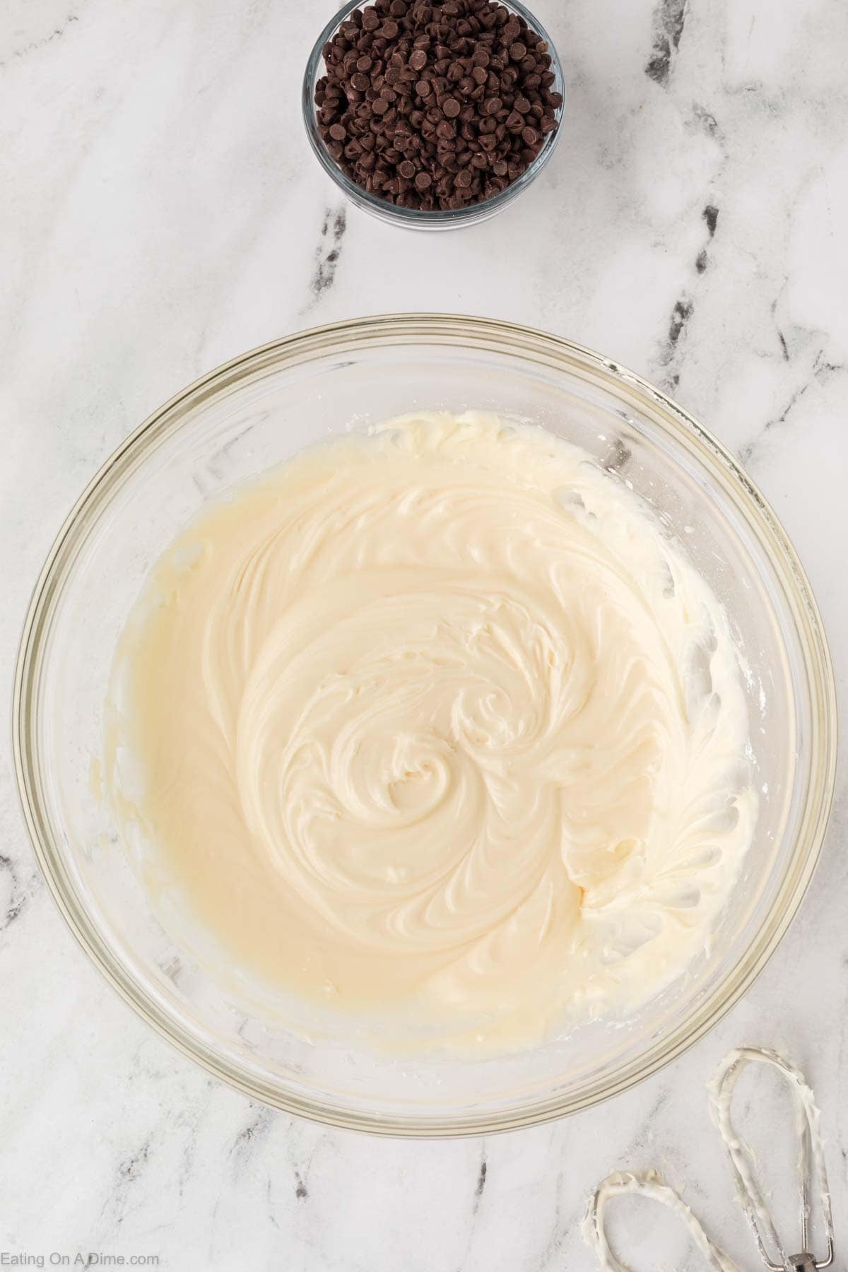 A glass bowl with creamy, swirled cheesecake batter for Red Velvet Poke Cake sits on a marble surface. Nearby are metal beaters with some batter and a small bowl filled with mini chocolate chips.