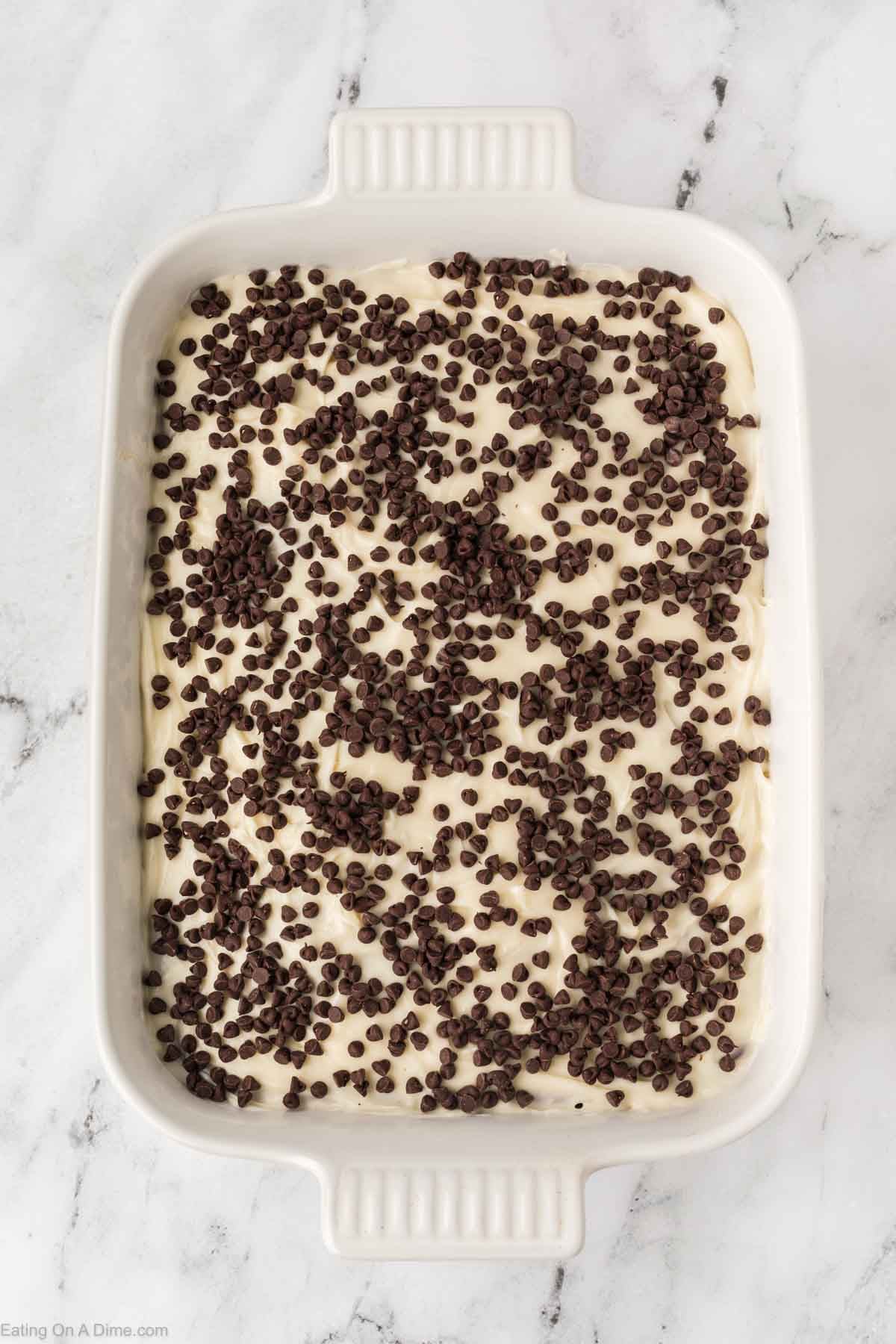 A white rectangular baking dish filled with creamy Red Velvet Poke Cake batter, topped with scattered mini chocolate chips, sits on a white marble surface.