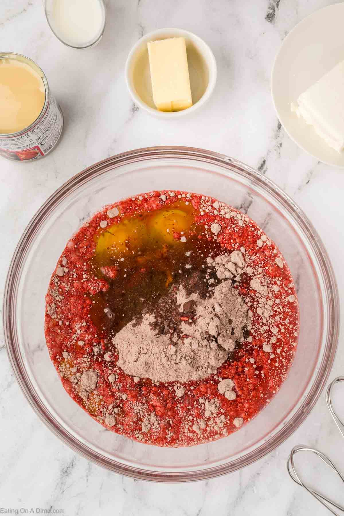 A glass mixing bowl with eggs, red cake mix, and cocoa powder sits on a white marble counter—perfect for making Red Velvet Poke Cake. Nearby are a stick of butter, cream cheese on a plate, a can of condensed milk, and an electric mixer.