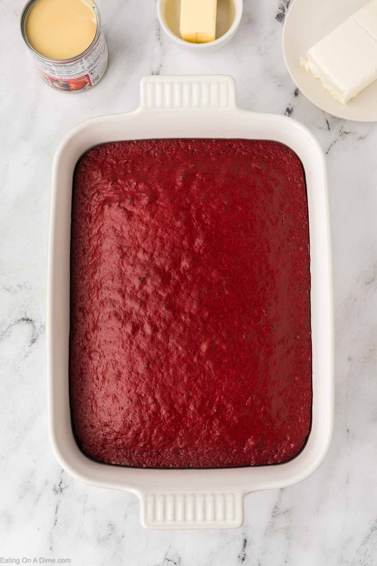 A rectangular white baking dish filled with a freshly baked Red Velvet Poke Cake sits on a marble surface, with butter, cream cheese, and a can of sweetened condensed milk nearby.