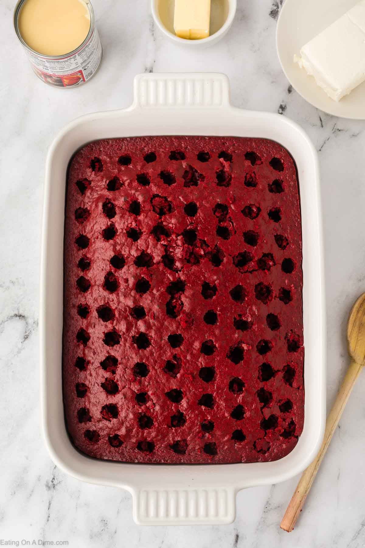 A rectangular white baking dish holds a Red Velvet Poke Cake with evenly spaced holes on top. Nearby are a wooden spoon, a can of sweetened condensed milk, and plates with butter and cream cheese.