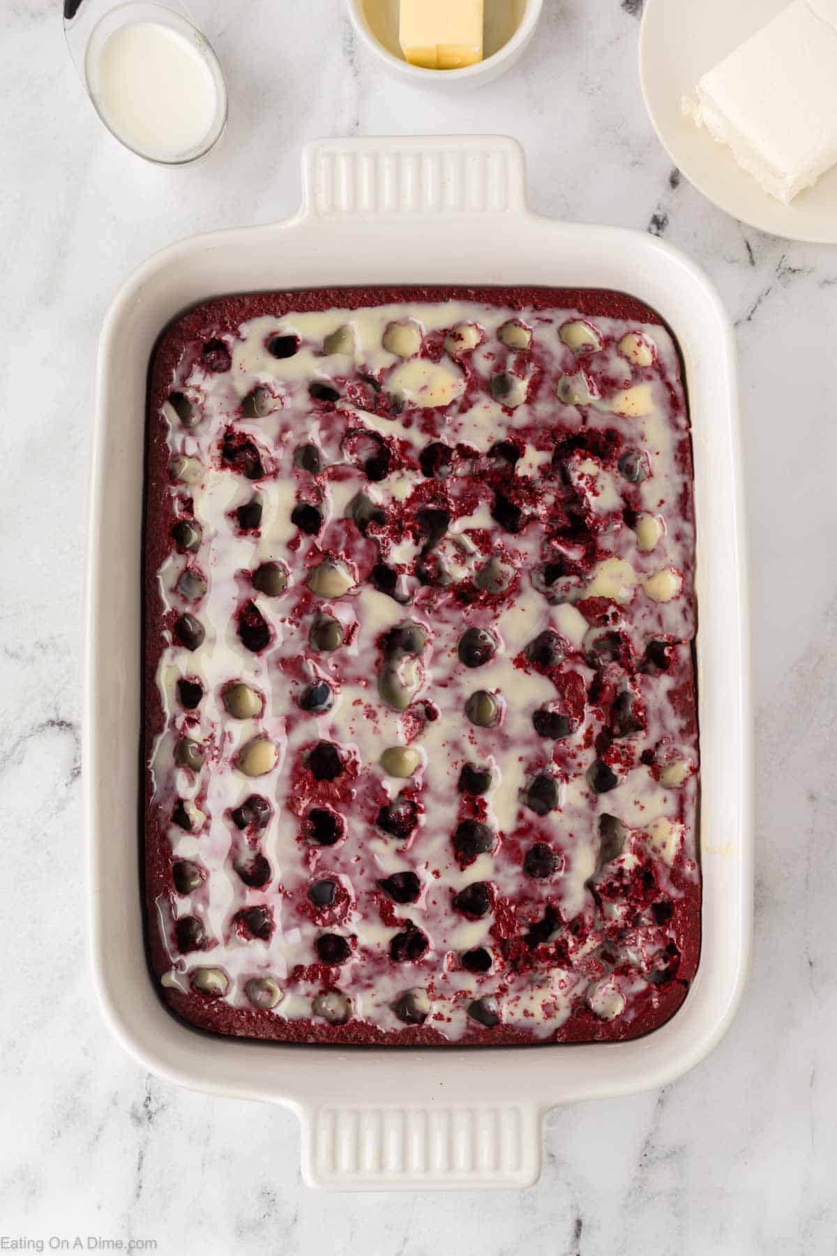 A rectangular white baking dish holds a Red Velvet Poke Cake topped with creamy white glaze, the glaze soaking into visible holes, all set on a marble countertop beside butter, milk, and cream cheese.