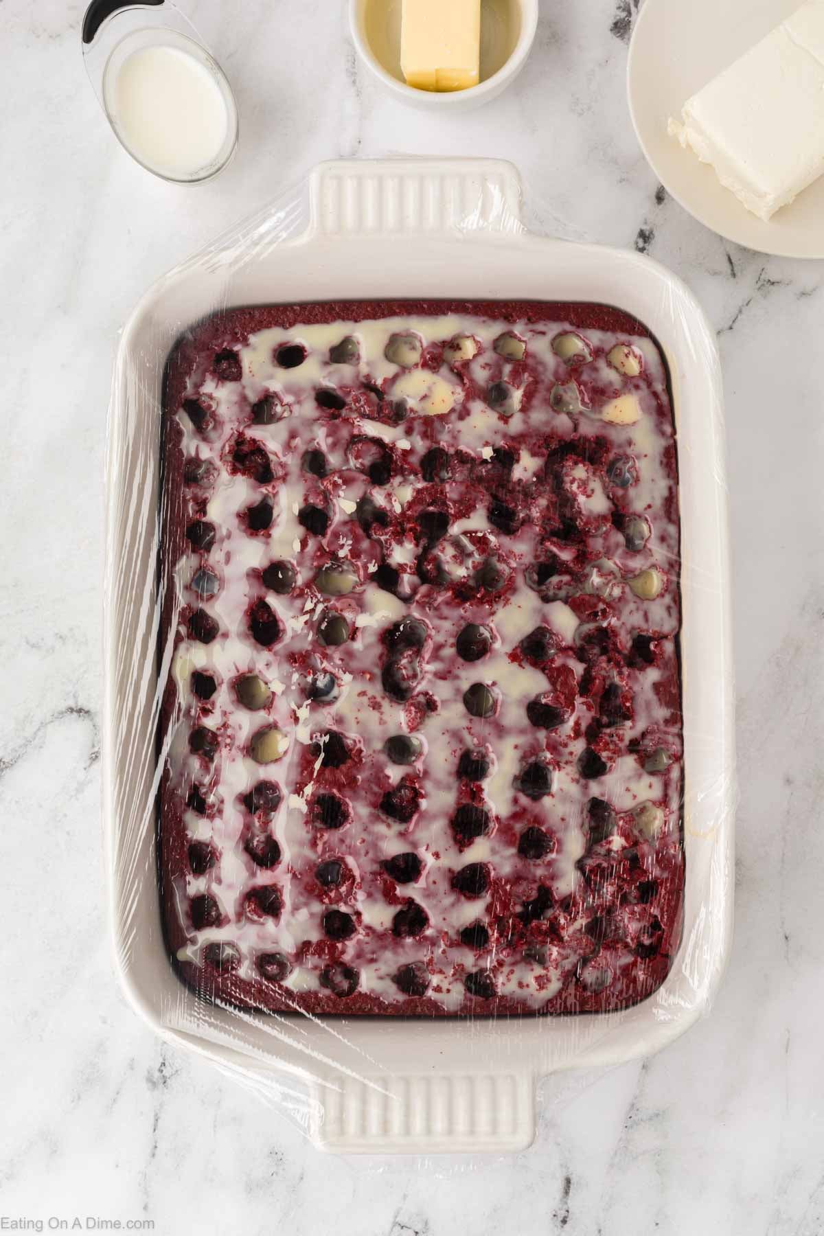 A rectangular baking dish filled with Red Velvet Poke Cake, drizzled with white glaze and covered with plastic wrap, sits on a marble surface. Nearby are a cup of cream, a stick of butter, and a plate with cream cheese.