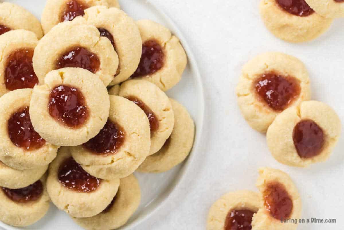 A plate piled with homemade thumbprint cookies filled with strawberry jam. On the white surface next to the plate, a few additional raspberry thumbprint cookies are scattered. The cookies have a golden-brown color and a distinct jam center.