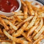 A plate of crispy golden air fryer French fries garnished with chopped herbs, served with a small bowl of ketchup in the background.
