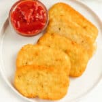 A white plate holds five golden-brown hash browns, crisp from an air fryer hashbrowns recipe, arranged in a row and served with a small glass bowl of chunky ketchup.