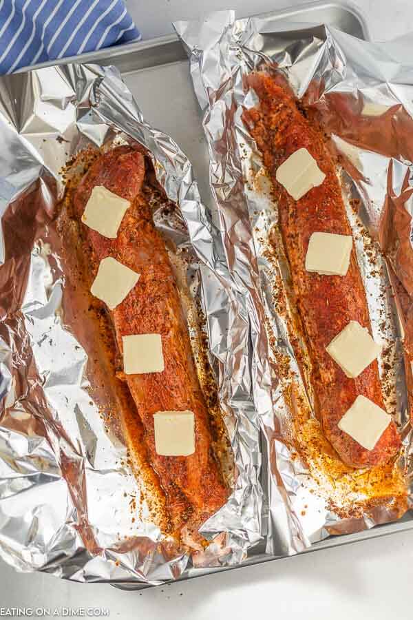 The raw pork tenderloin placed on a piece of foil, topped with oil, seasonings and slabs of butter 