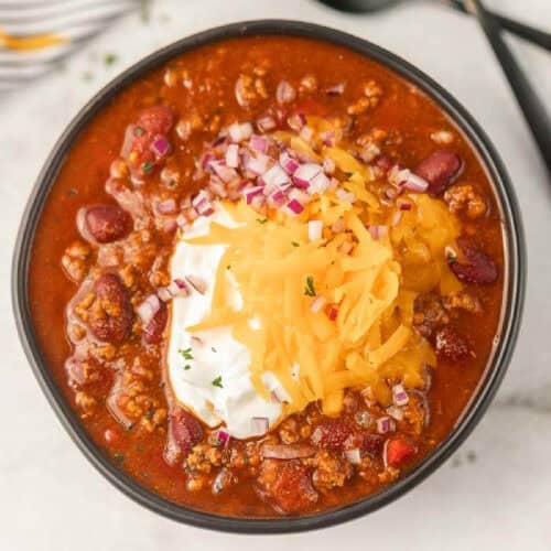 A bowl of Crockpot Cowboy Chili topped with shredded cheddar cheese, a dollop of sour cream, and chopped red onions, featuring ground meat, beans, and tomato pieces in a rich, red sauce.