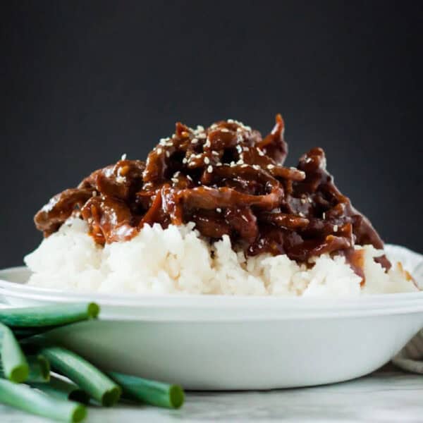 A white plate filled with steamed white rice topped with saucy Slow Cooker Mongolian Beef, garnished with sesame seeds and served with green onions on the side.