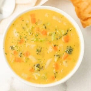A bowl of creamy vegetable soup recipe with visible chunks of carrots, broccoli, potatoes, and herbs. A piece of bread is partially visible on the side.