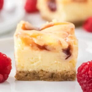 A close-up of raspberry cheesecake bars with a graham cracker crust and a swirl of raspberry sauce on top, surrounded by fresh raspberries on a white plate.