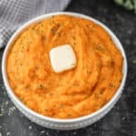 A bowl of creamy Instant Pot mashed sweet potatoes topped with a pat of butter and fresh herbs, placed on a dark surface next to a checkered cloth.