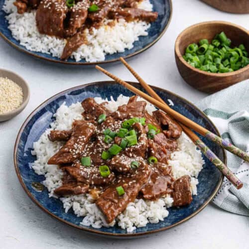 A blue plate filled with white rice topped with saucy Crock Pot Sesame Beef strips, garnished with chopped green onions and sesame seeds. Chopsticks rest on the plate, with bowls of green onions and sesame seeds nearby.
