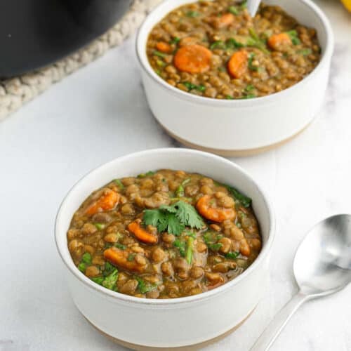 Two white bowls filled with curry lentil soup containing carrots and garnished with fresh cilantro sit on a white surface next to a spoon.