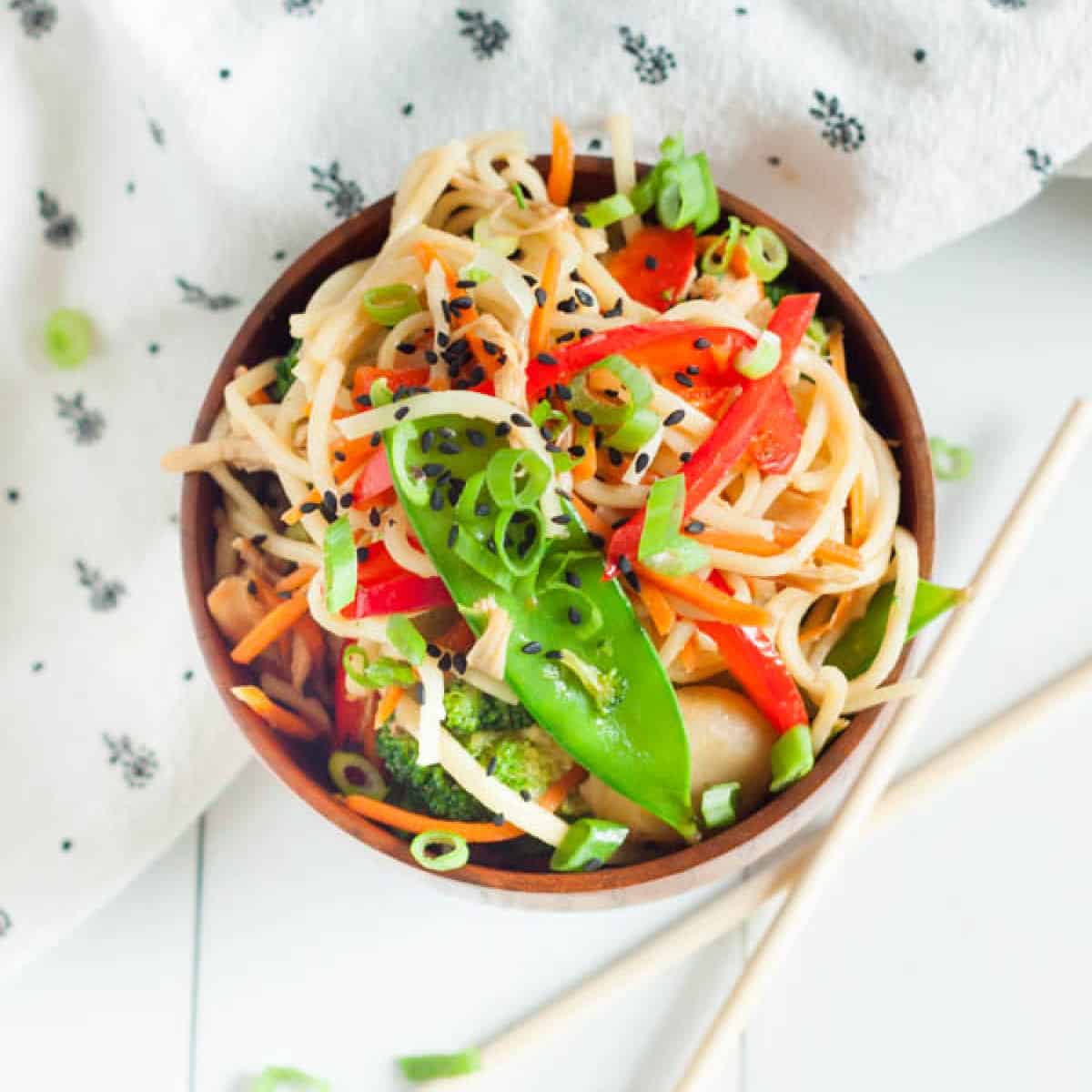 A colorful bowl of Asian noodle salad with sliced red bell peppers, snow peas, shredded carrots, and black sesame seeds sits enticingly next to a patterned napkin. This dish, reminiscent of a vibrant crock pot chicken chow mein recipe, is accompanied by chopsticks resting aside.