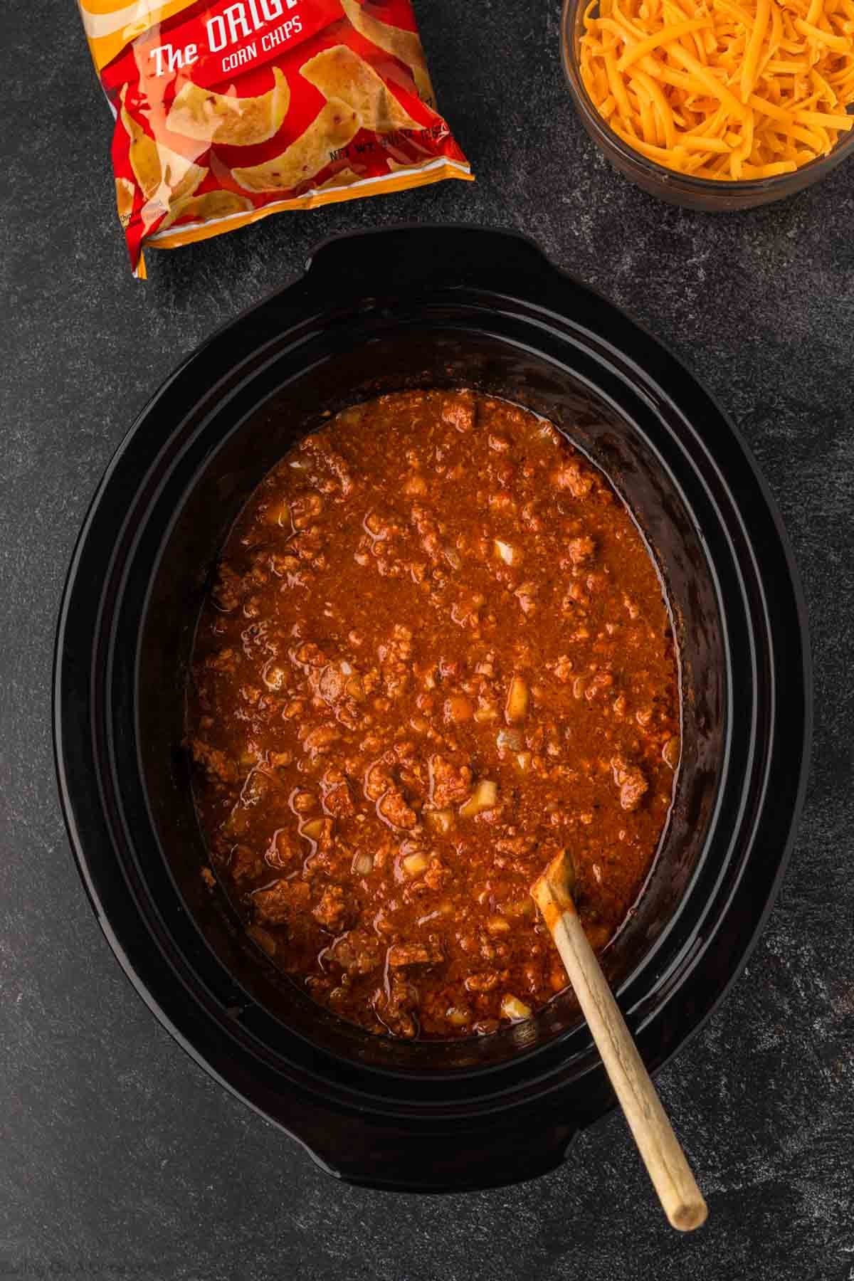 A Crock Pot Frito Pie simmers in a slow cooker with chili and a wooden spoon on a dark surface, next to a bowl of shredded cheddar cheese and a bag of corn chips.
