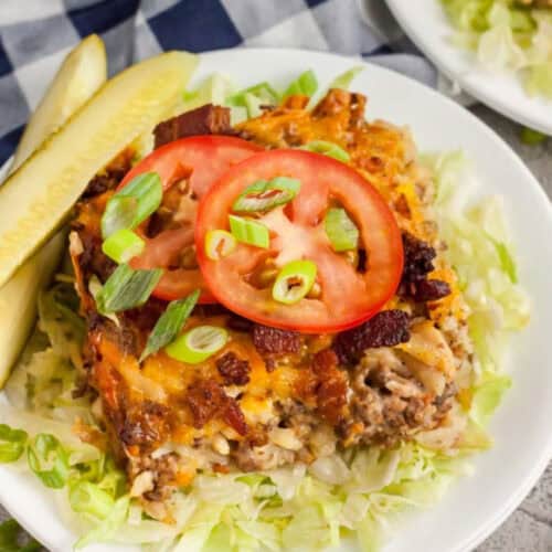 Bacon cheeseburger casserole topped with sliced tomatoes, green onions, and melted cheese, served on a bed of shredded lettuce with crispy bacon and two pickle spears on the side.