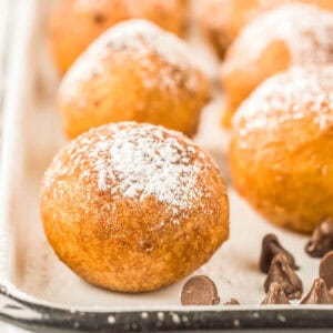 Close-up of golden brown Fried Cookie Dough balls dusted with powdered sugar, resting on a white tray with chocolate chips scattered nearby.