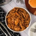 A white bowl filled with shredded instant pot pork tacos sits on a countertop beside a black checkered towel, metal tongs, forks, a sandwich bun, and white plates.