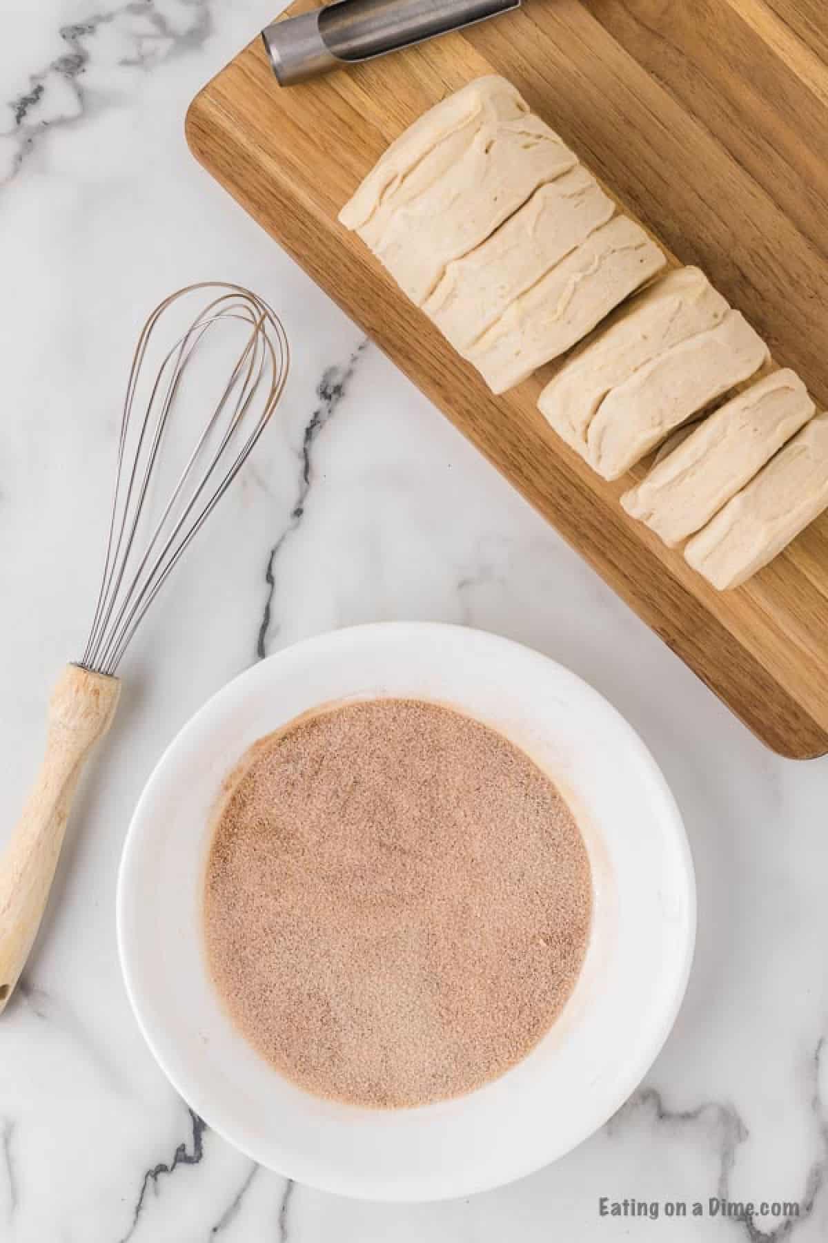 A bowl of cinnamon and sugar mixture with a cutting board with biscuit dough stacked.