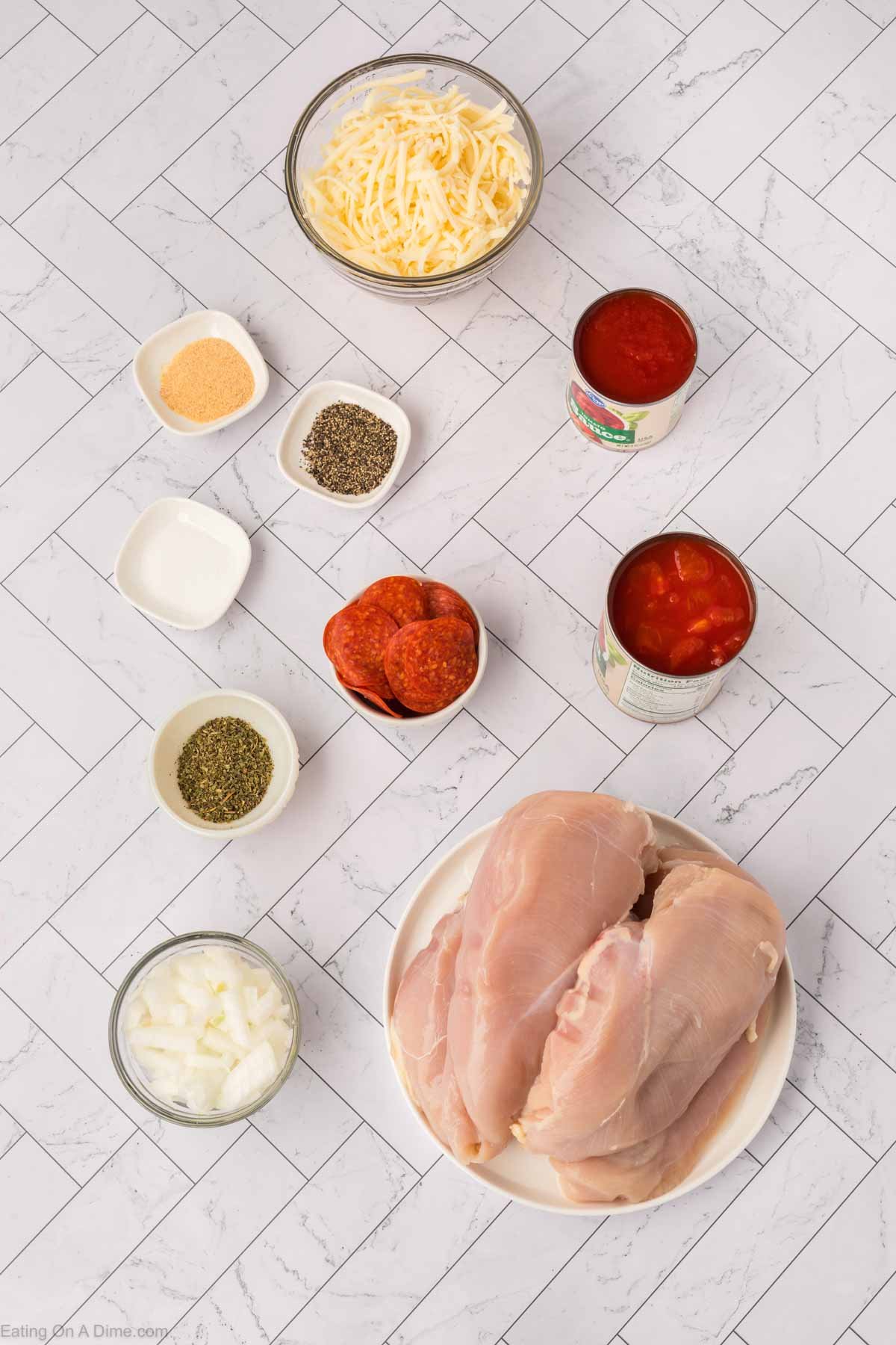 A flatlay photo of raw chicken breasts on a plate, sliced onions, spices in small bowls, shredded cheese, pepperoni slices, a can of tomato sauce, and a can of diced tomatoes—perfect ingredients for a Low Carb Pizza Chicken Recipe—on a white herringbone-patterned surface.
