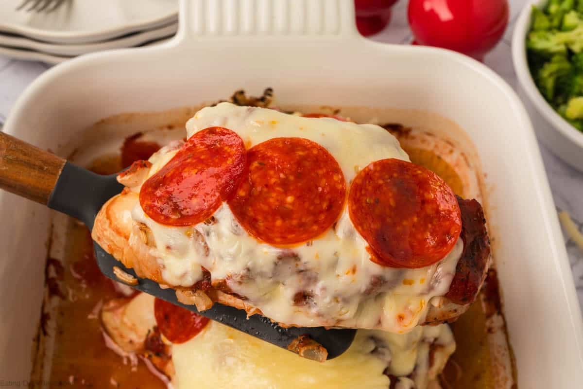 A spatula lifts a piece of Low Carb Pizza Chicken Recipe—baked chicken breast topped with melted cheese and pepperoni slices—from a white baking dish. Plates, red seasoning shakers, and broccoli are visible in the background.