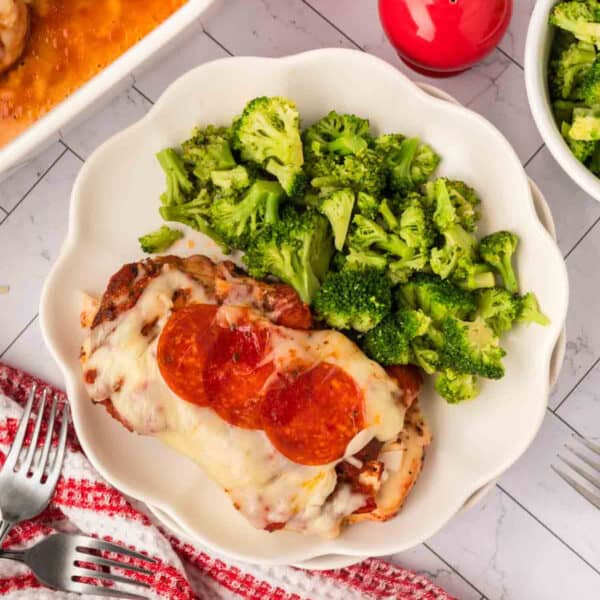 A white plate with Low Carb Pizza Chicken Recipe—baked chicken breast topped with melted cheese and pepperoni slices, served alongside steamed broccoli. A red and white striped towel, forks, and a dish of extra broccoli are nearby.