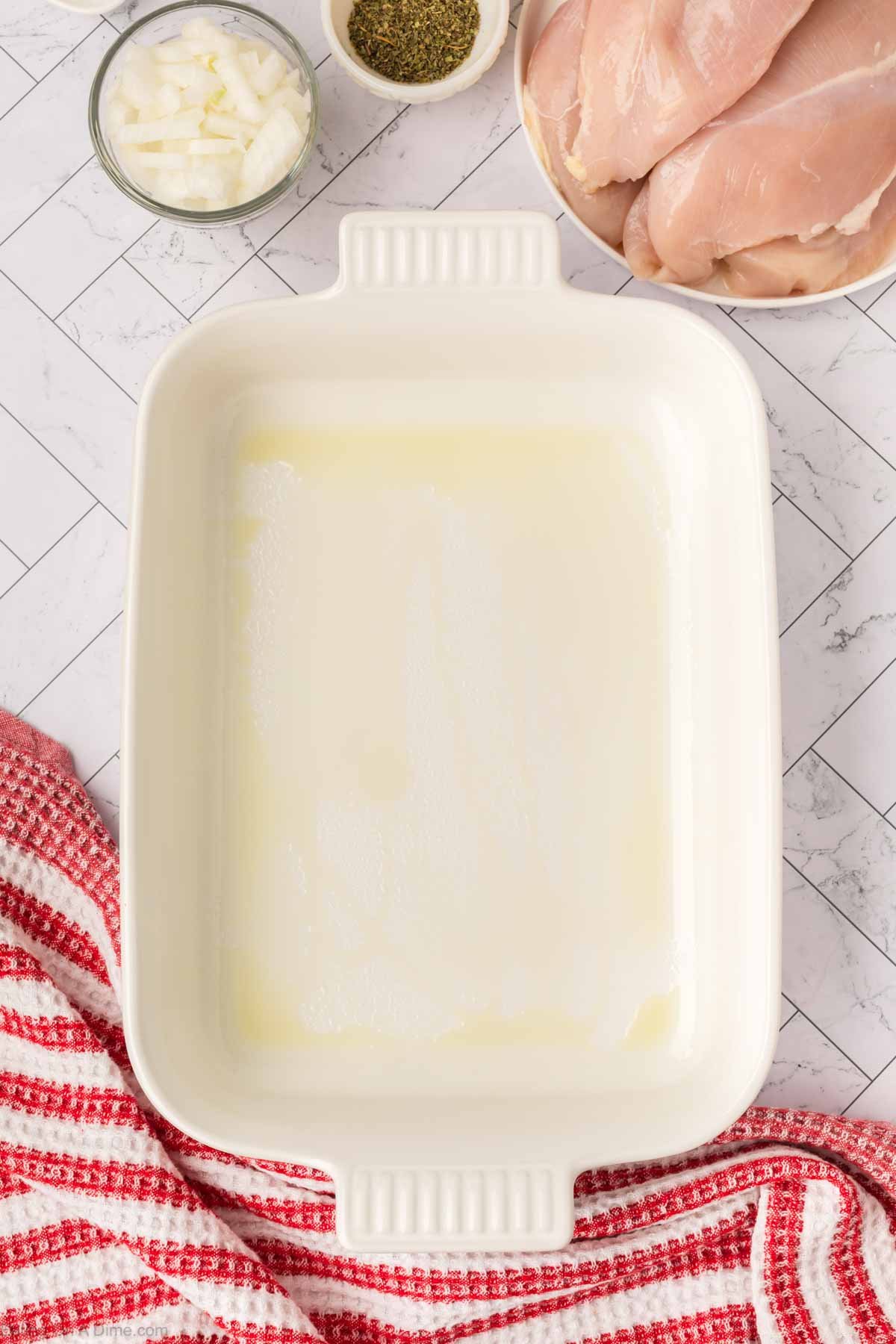 A greased white rectangular baking dish sits on a marble countertop, ready for this Low Carb Pizza Chicken Recipe. Surrounding it are bowls of chopped onions, dried herbs, raw chicken breasts, and a red-and-white striped towel.