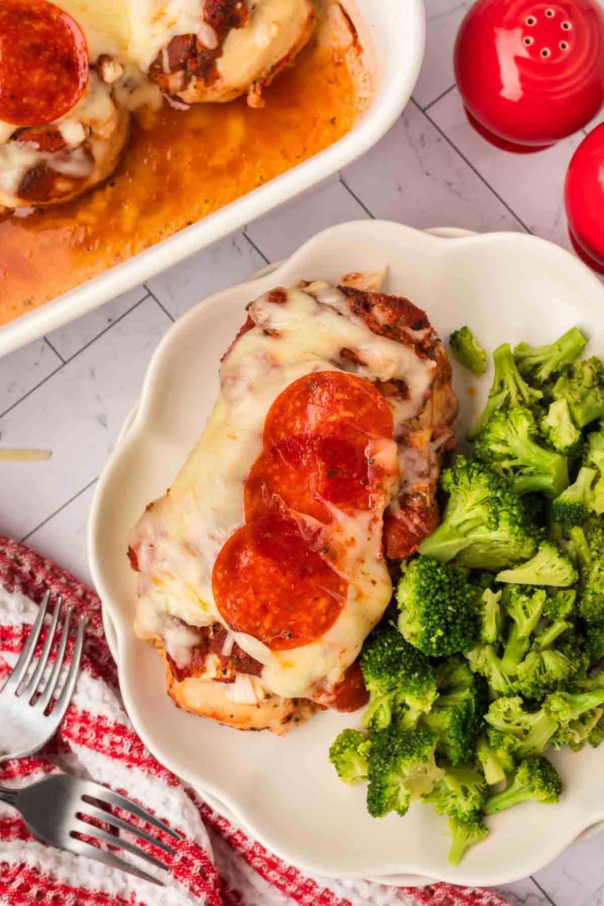 A plate with Low Carb Pizza Chicken Recipe: baked chicken topped with melted cheese and pepperoni slices, served with steamed broccoli. A baking dish with more chicken, two red salt and pepper shakers, and a red-striped towel are nearby.