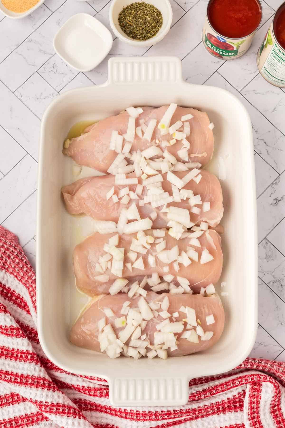 Four raw chicken breasts topped with chopped onions are arranged in a white baking dish for this Low Carb Pizza Chicken Recipe. The dish sits on a marble countertop near a red kitchen towel, herbs, and cans of tomatoes.
