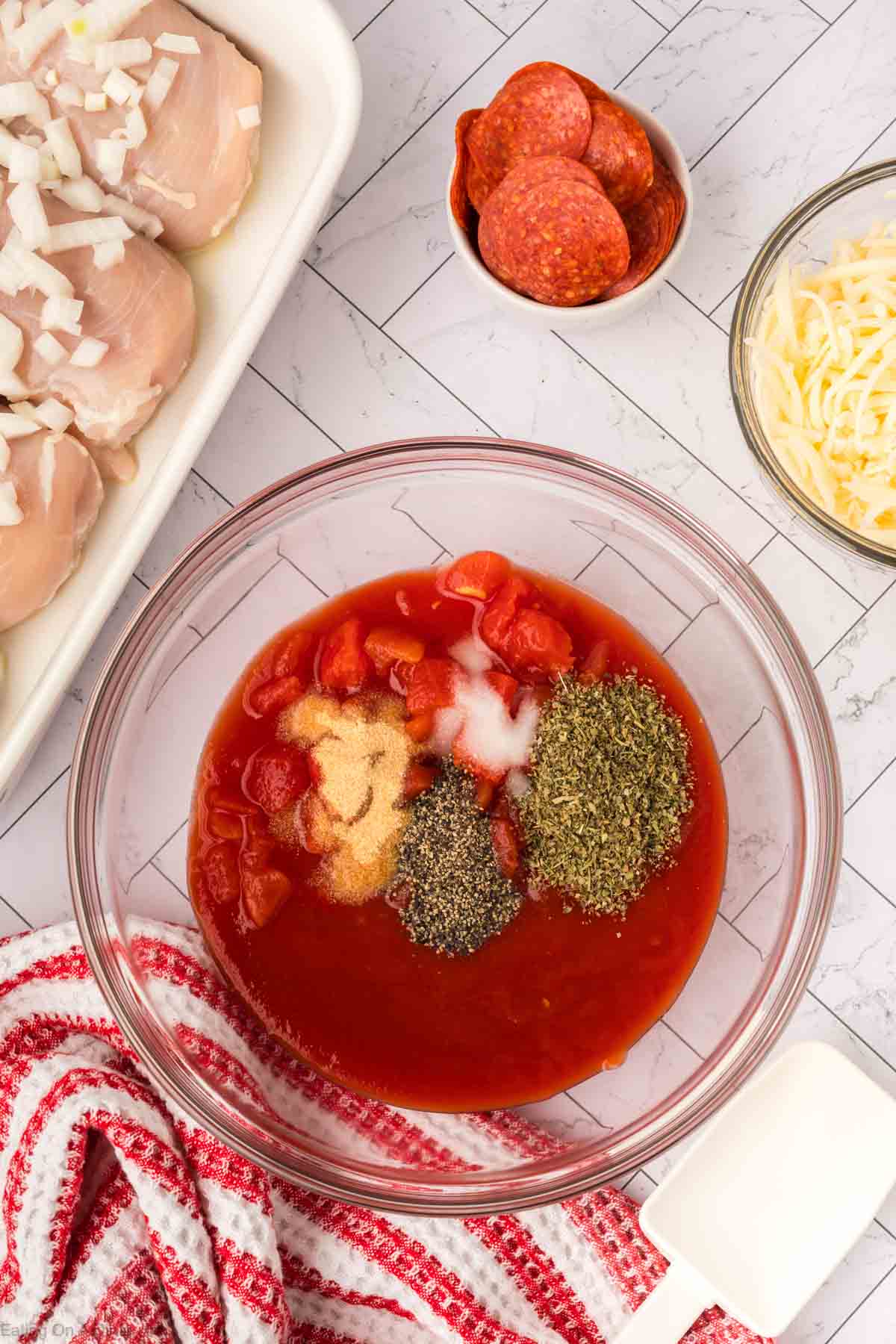 A glass bowl with tomato sauce, diced tomatoes, herbs, and spices sits on a counter. Nearby are raw chicken breasts for this Low Carb Pizza Chicken Recipe, along with onions, pepperoni slices, shredded cheese, and a red-striped towel.